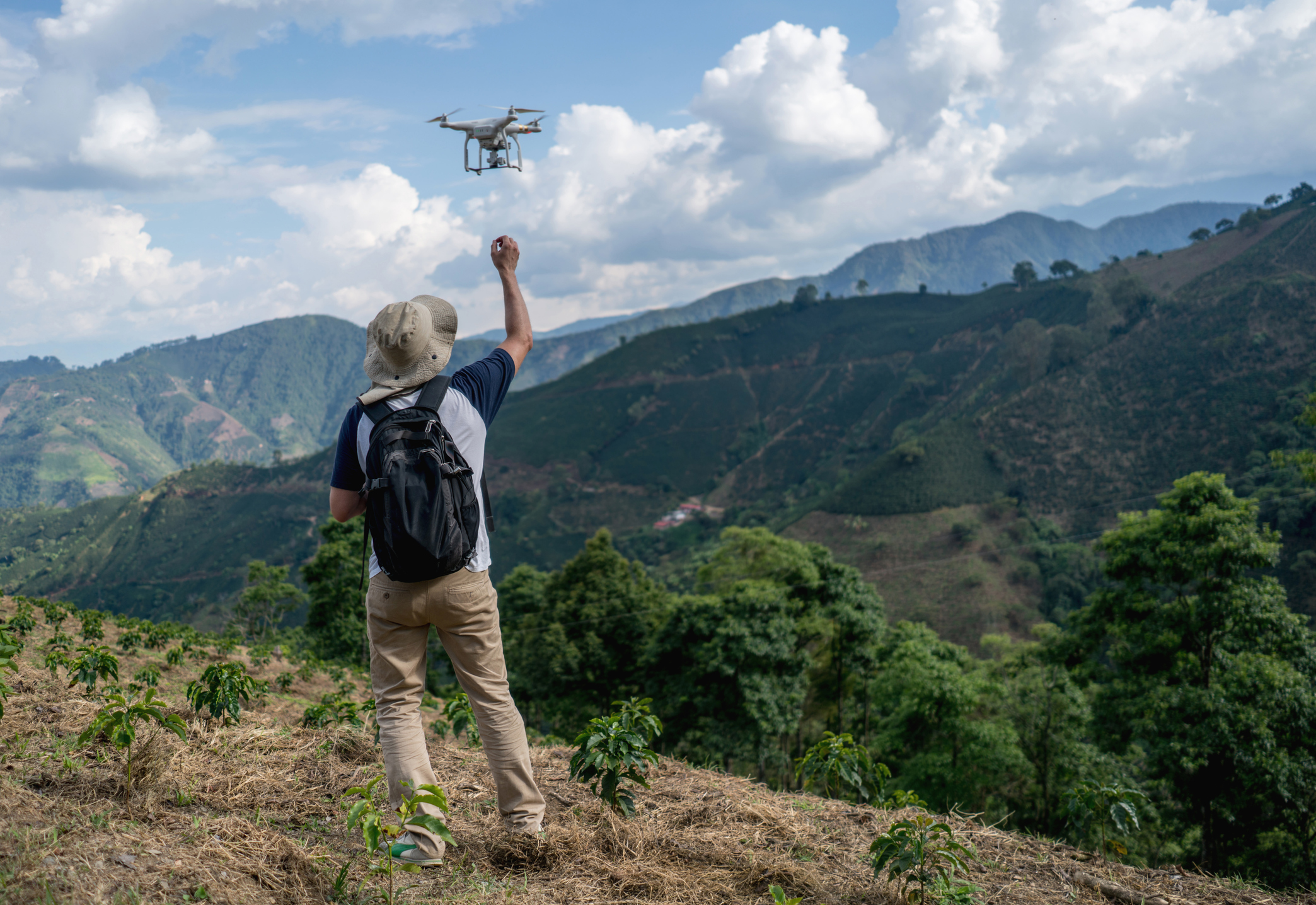 A man in a wide-brimmed hat and backpack flies a drone over lush, rolling hills and farmland under a partly cloudy sky